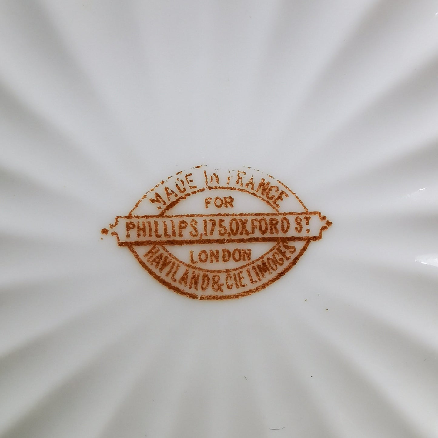 Close-up of the inside bottom of a white fluted ceramic dish, showing a brown stamp that reads 'MADE IN FRANCE FOR PHILLIPS 175 OXFORD SHILLINGTON & CO LONDON'.