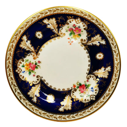 A round porcelain plate with a classic Royal Navy blue border, decorated with gold leaf patterns, white floral motifs, and colorful pink and yellow flowers, finished with a gold rim.