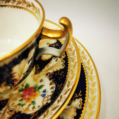 An ornate porcelain teacup and saucer set with a navy blue and gold baroque-style pattern, featuring a white interior and a hand-painted floral motif.