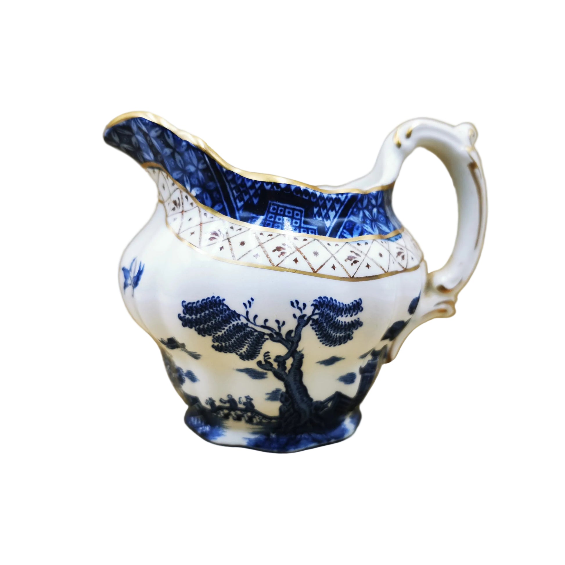 A vintage blue and white porcelain creamer pitcher with gold trim, featuring a landscape design of trees and mountains, set against a tan cardboard background.