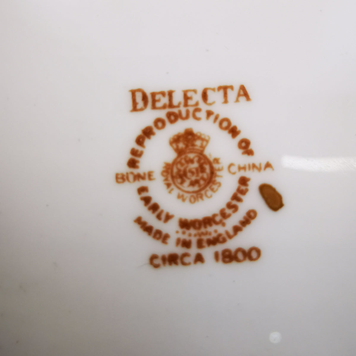 Close-up of a white ceramic surface printed with a brown reproduction mark reading 'DELECTA' and 'Reproduction of Early Worcester Made in England circa 1800' with a crest.