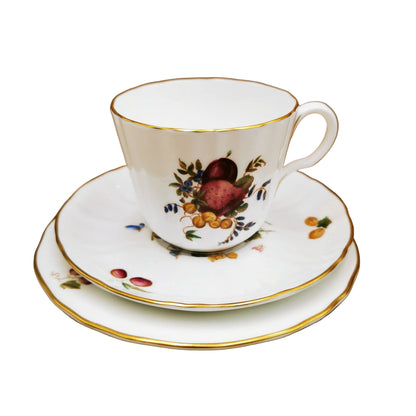 A white porcelain teacup and saucer set with gold trim and a hand-painted fruit motif, displayed on a matching saucer against a brown background.