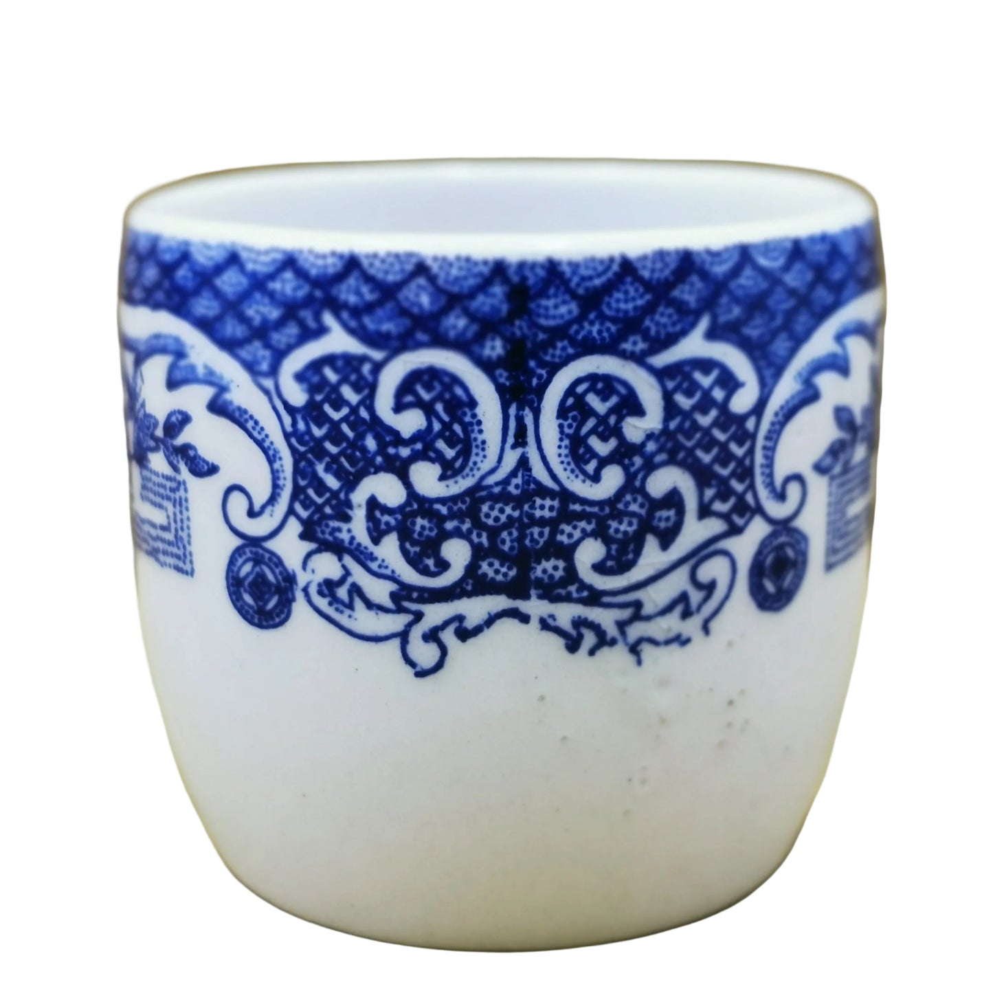 A small white ceramic cup or tumbler with a blue ornamental scroll pattern around the upper half, photographed against a tan cardboard-like background.