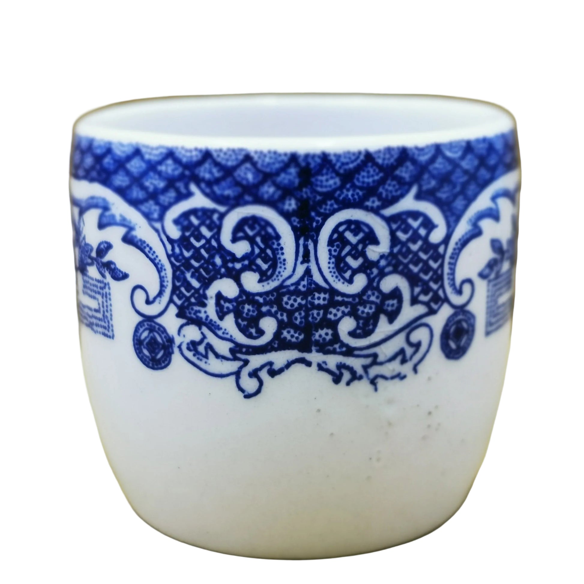 A small white ceramic cup or tumbler with a blue ornamental scroll pattern around the upper half, photographed against a tan cardboard-like background.