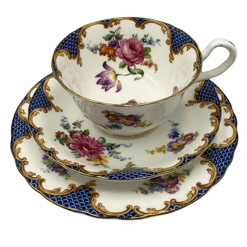 A vintage porcelain teacup and saucer set with a floral design, featuring pink roses, purple tulips, blue and gold patterns, and a white handle, set against a plain light gray background.