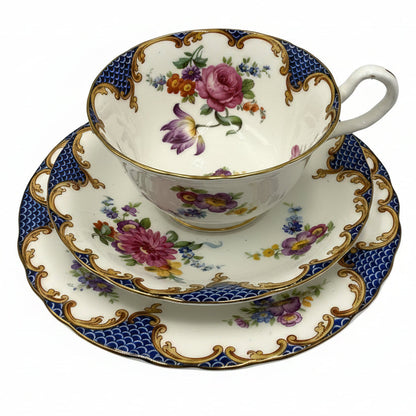 A vintage porcelain teacup and saucer set with a floral design, featuring pink roses, purple tulips, blue and gold patterns, and a white handle, set against a plain light gray background.