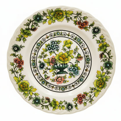 A white ceramic plate with a hand-painted floral design featuring green leaves, yellow flowers, blue blossoms, and red accents, set against a neutral gray background.