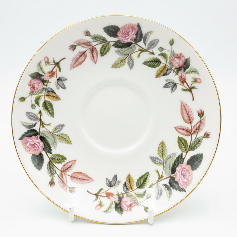 A white porcelain saucer decorated with a delicate floral border of pink roses, green and pink leaves, and a thin gold rim, displayed on a cardboard backing.