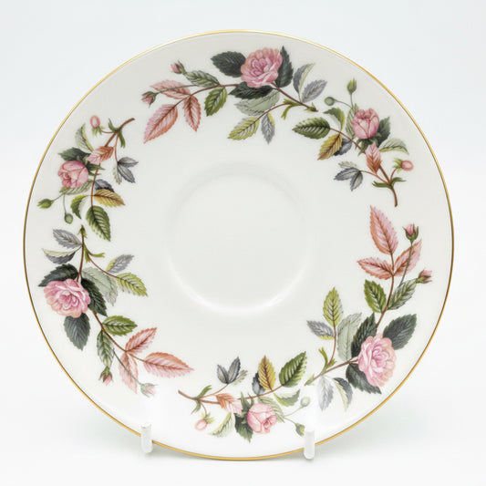A white porcelain saucer decorated with a delicate floral border of pink roses, green and pink leaves, and a thin gold rim, displayed on a cardboard backing.
