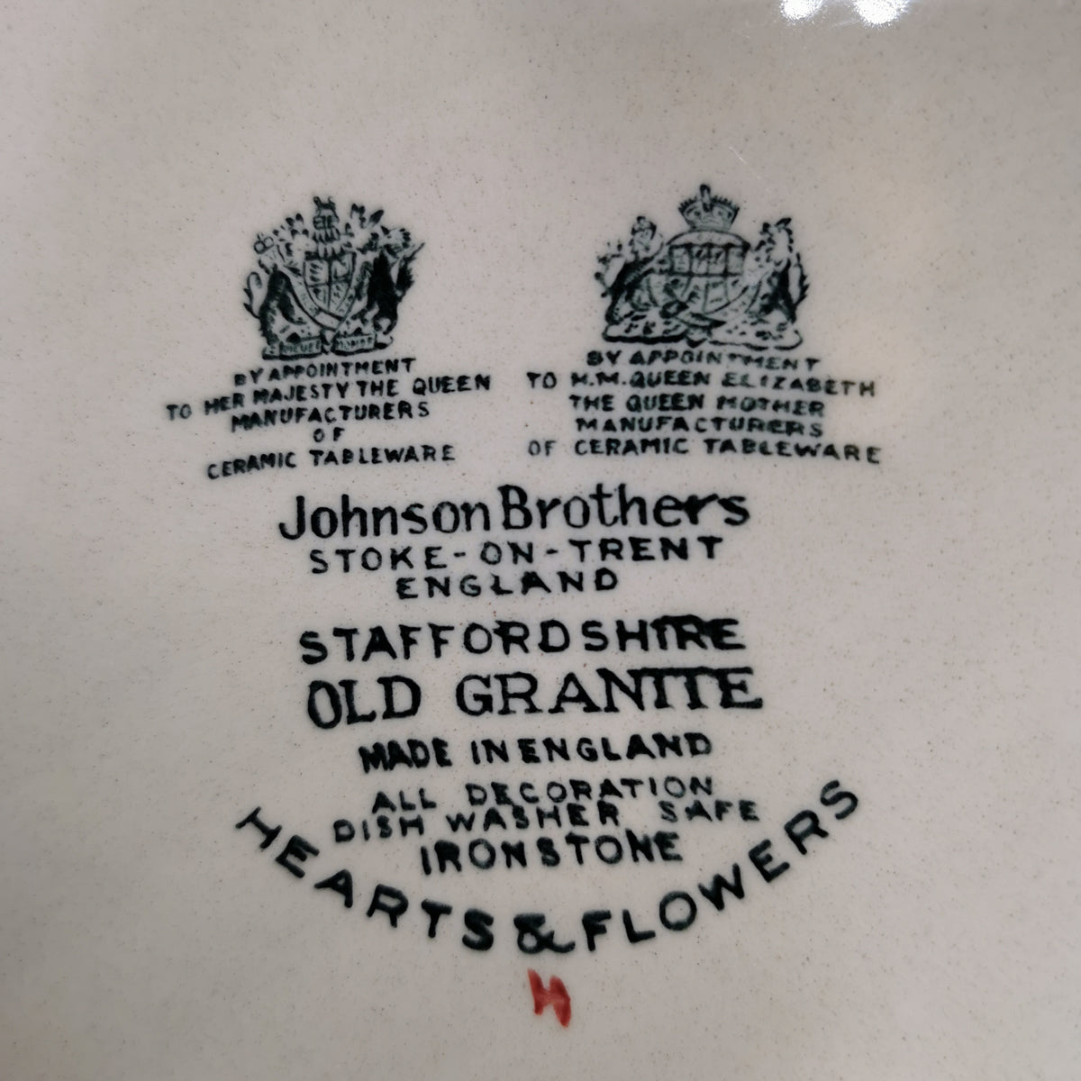 Close-up of a white ceramic plate or dish printed with black text and two crest emblems. Visible text includes 'Johnson Brothers', 'Stoke-on-Trent England', 'Staffordshire Old Granite', 'Made in England', and 'Hearts & Flowers'.