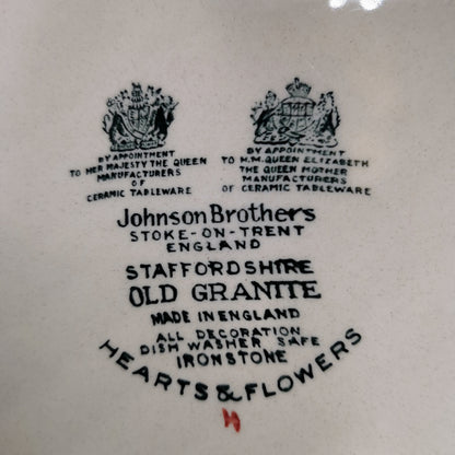 Close-up of a white ceramic plate or dish printed with black text and two crest emblems. Visible text includes 'Johnson Brothers', 'Stoke-on-Trent England', 'Staffordshire Old Granite', 'Made in England', and 'Hearts & Flowers'.