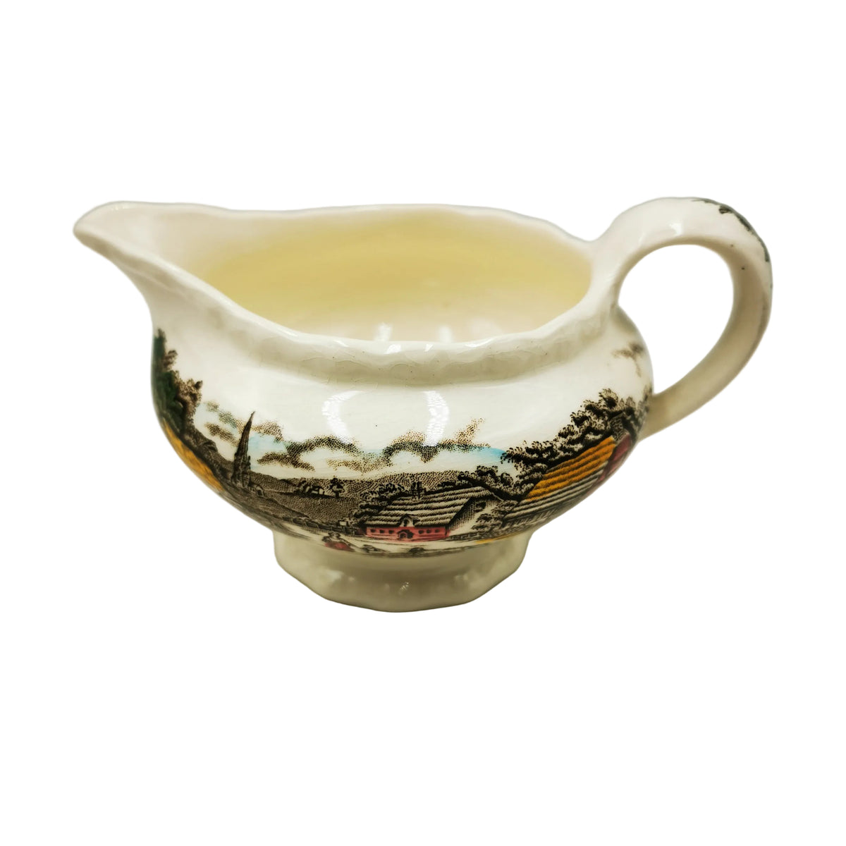 An antique cream-colored porcelain gravy boat with a hand-painted landscape design featuring houses, trees, and mountains in black, brown, yellow, and red, set against a neutral gray background.