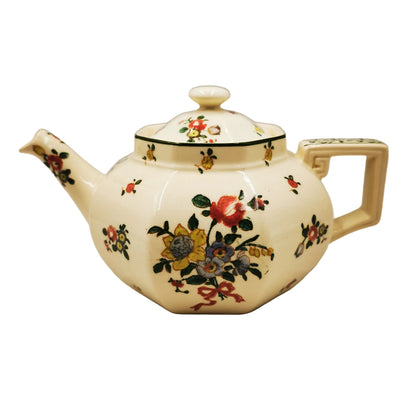 An antique-style ceramic teapot with a cream-colored body, decorated with a colorful floral pattern and a green band around the neck, set against a plain light gray background.