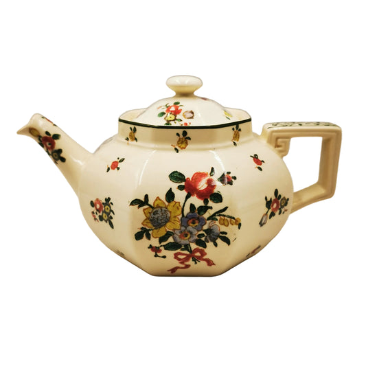An antique-style ceramic teapot with a cream-colored body, decorated with a colorful floral pattern and a green band around the neck, set against a plain light gray background.