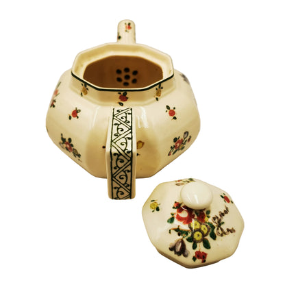 An antique-style ceramic octagonal teapot with a matching lid, decorated with a cream glaze, small red and yellow floral motifs, and black geometric patterns on the handle and neck.