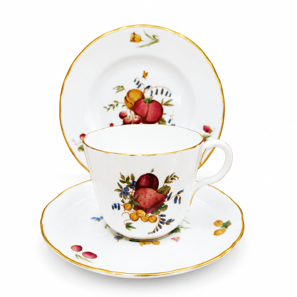 A white porcelain teacup and saucer set with gold trim and a hand-painted fruit motif, shown with a matching plate against a brown background.