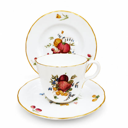 A white porcelain teacup and saucer set with gold trim and a hand-painted fruit motif, shown with a matching plate against a brown background.