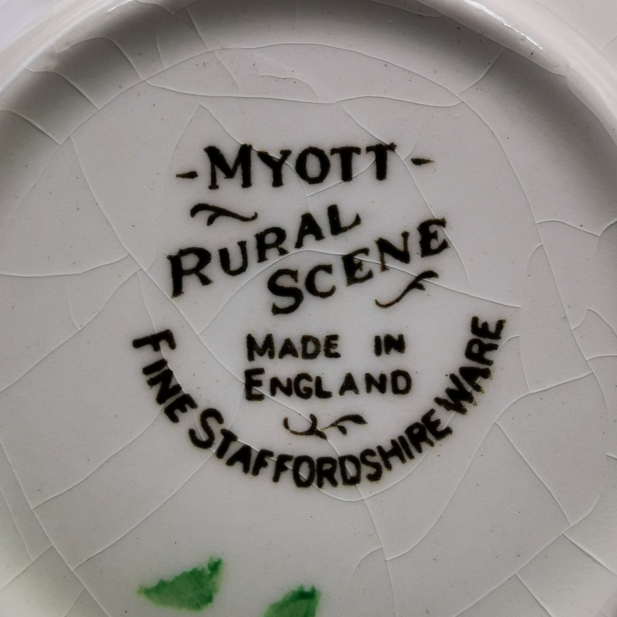 Close-up of the inside bottom of a white ceramic plate or dish with visible crackle glaze. Black printed text reads “MYOTT Rural Scene Made in England” and “FINE STAFFORDSHIRE WARE,” with a few small green marks near the bottom edge.