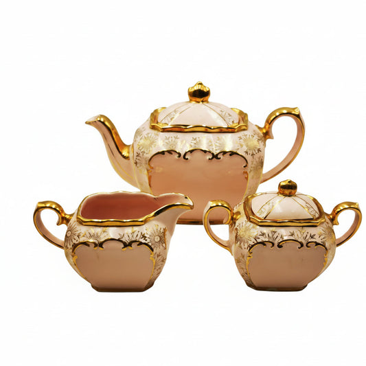 A three-piece ceramic tea and coffee set consisting of a teapot, a creamer, and a small lidded sugar bowl, all in a pale pink color with gold trim and floral detailing, set against a neutral light gray background.