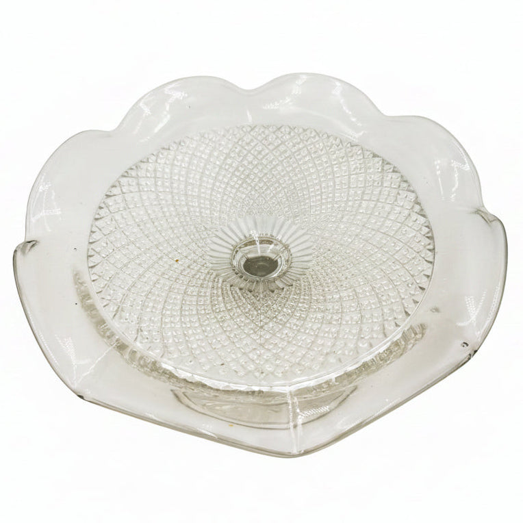 A clear glass pedestal plate with a scalloped rim and a textured, beaded circular pattern on the top, photographed on a neutral light gray background.