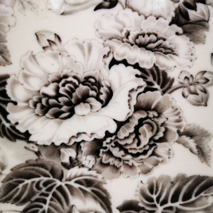 Close-up of a white ceramic surface decorated with a detailed black and gray floral pattern featuring peony-like blossoms and leaves.